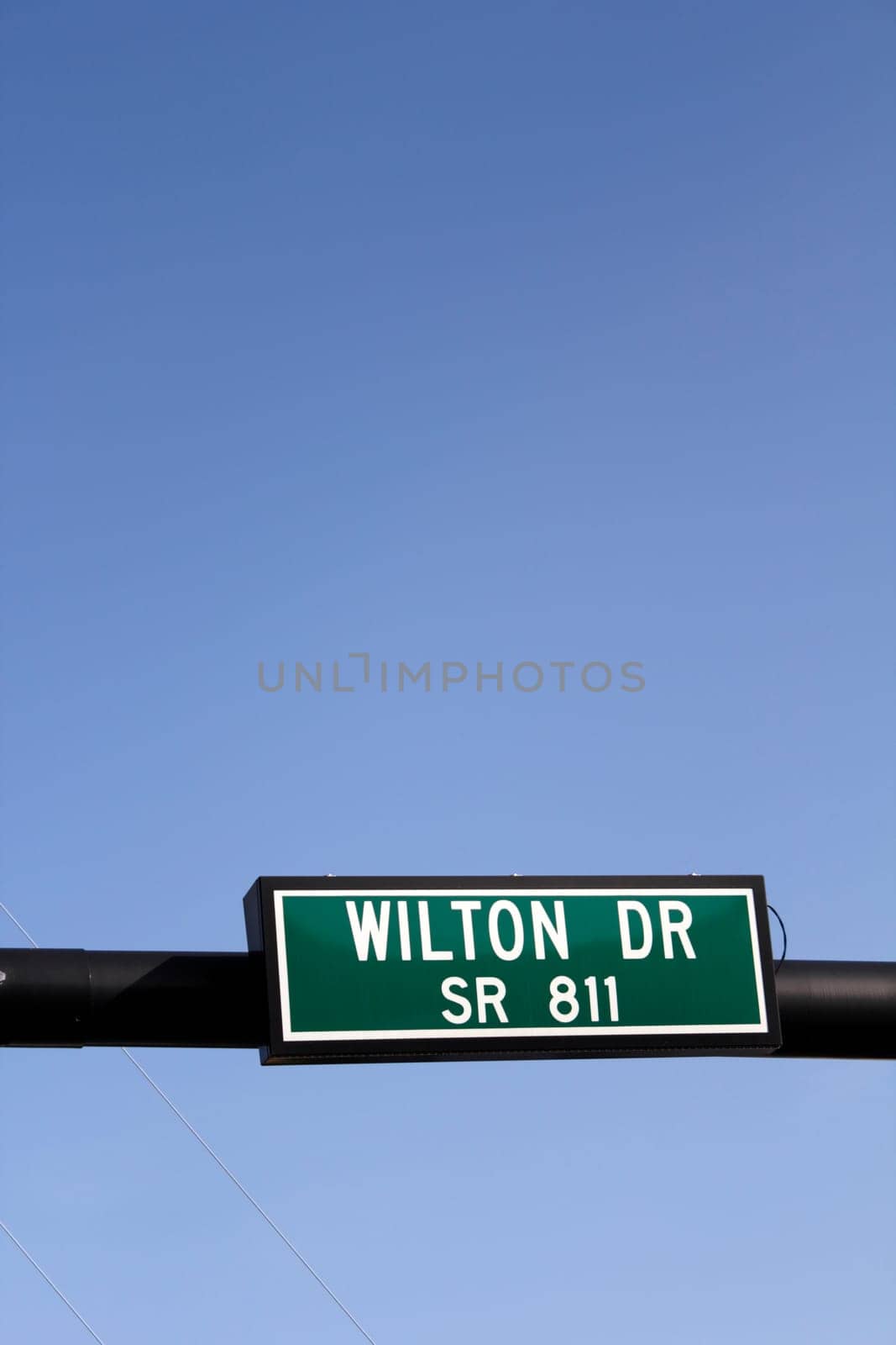 Rectangle white green and black street sign hanging from a pole on a sunny blue sky day in Wilton Manors, Florida..
