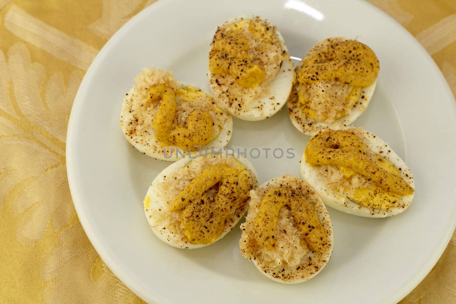 Three prepared boiled eggs halved with yolk prepared with deviled egg mixture and placed on a round white plate with a yellow gold floral place mat background..