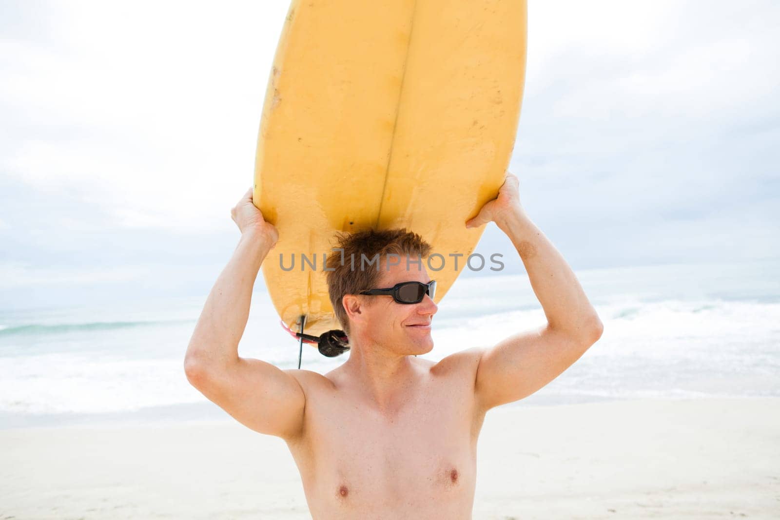 Smiling male surfer resting surfboard on head at beach with ocean and sky in background