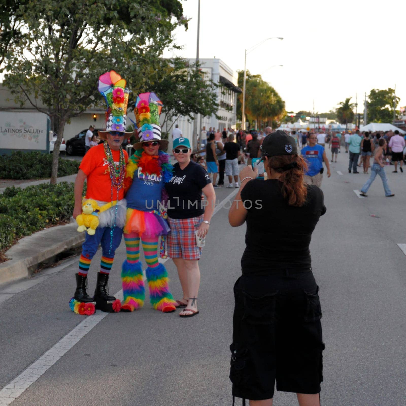 WILTON MANORS, FLORIDA - JUNE 22, 2013: Two adult people in colorful costumes with big hats pose next to a gay pride celebration attendee while another person makes a photograph on a smart phone.