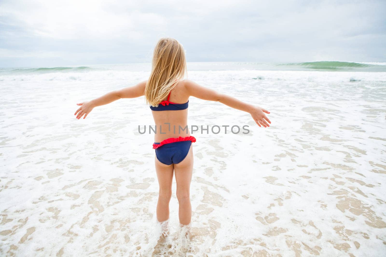 Cute young girl standing in water at beach facing the ocean with arms raised