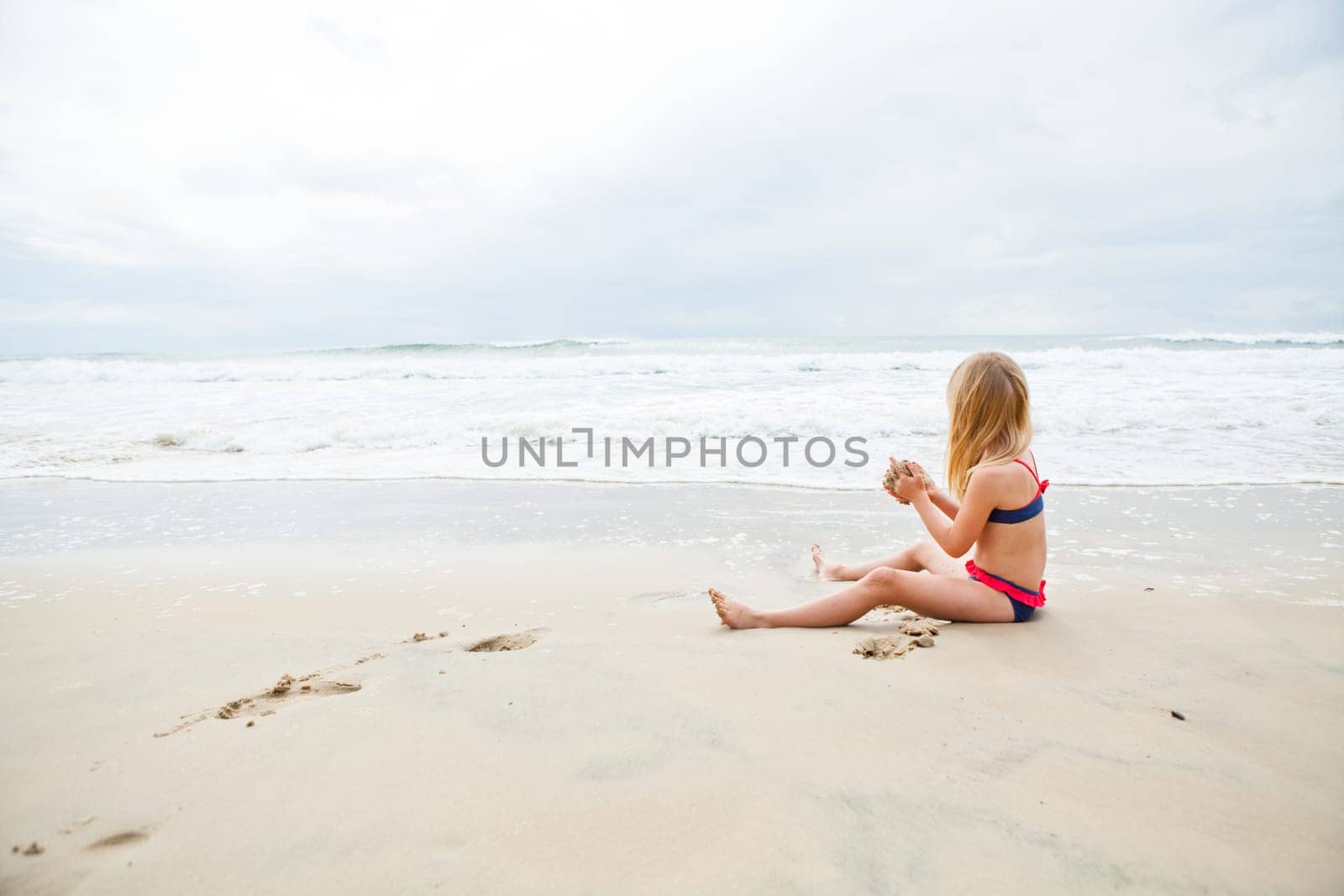 Young blond girl sitting on sand and playing at beach with ocean in background