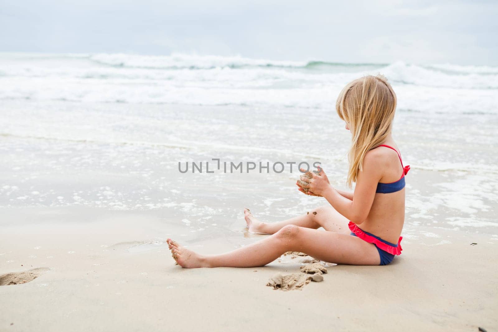 Cute young girl sitting on sand and playing at beach with ocean in background