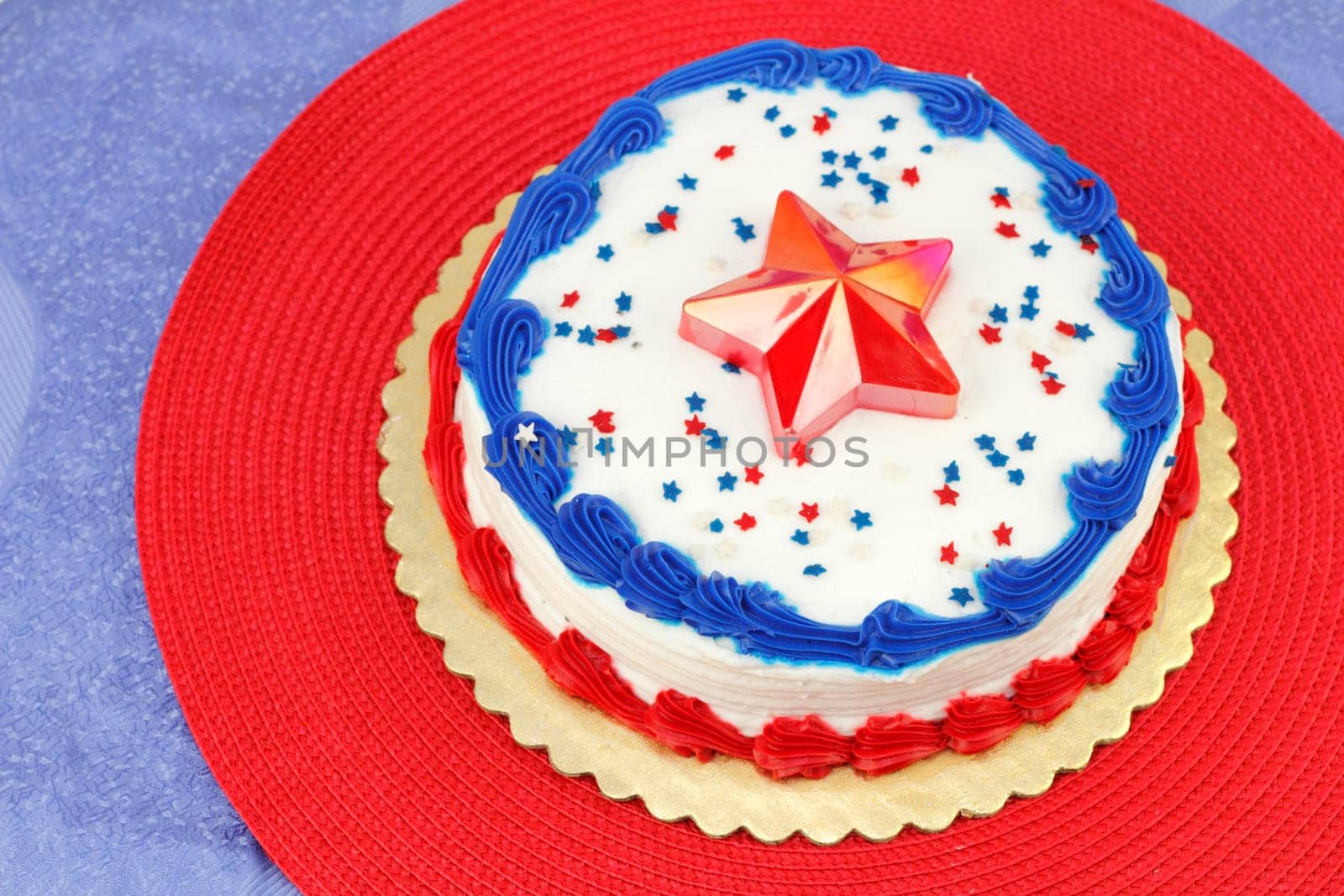 Round Independence Day holiday cake decorated with white frosting, red and blue ribbons, edible stars and a red plastic star on the center top. The cake is on a gold base, then red and blue placemats.