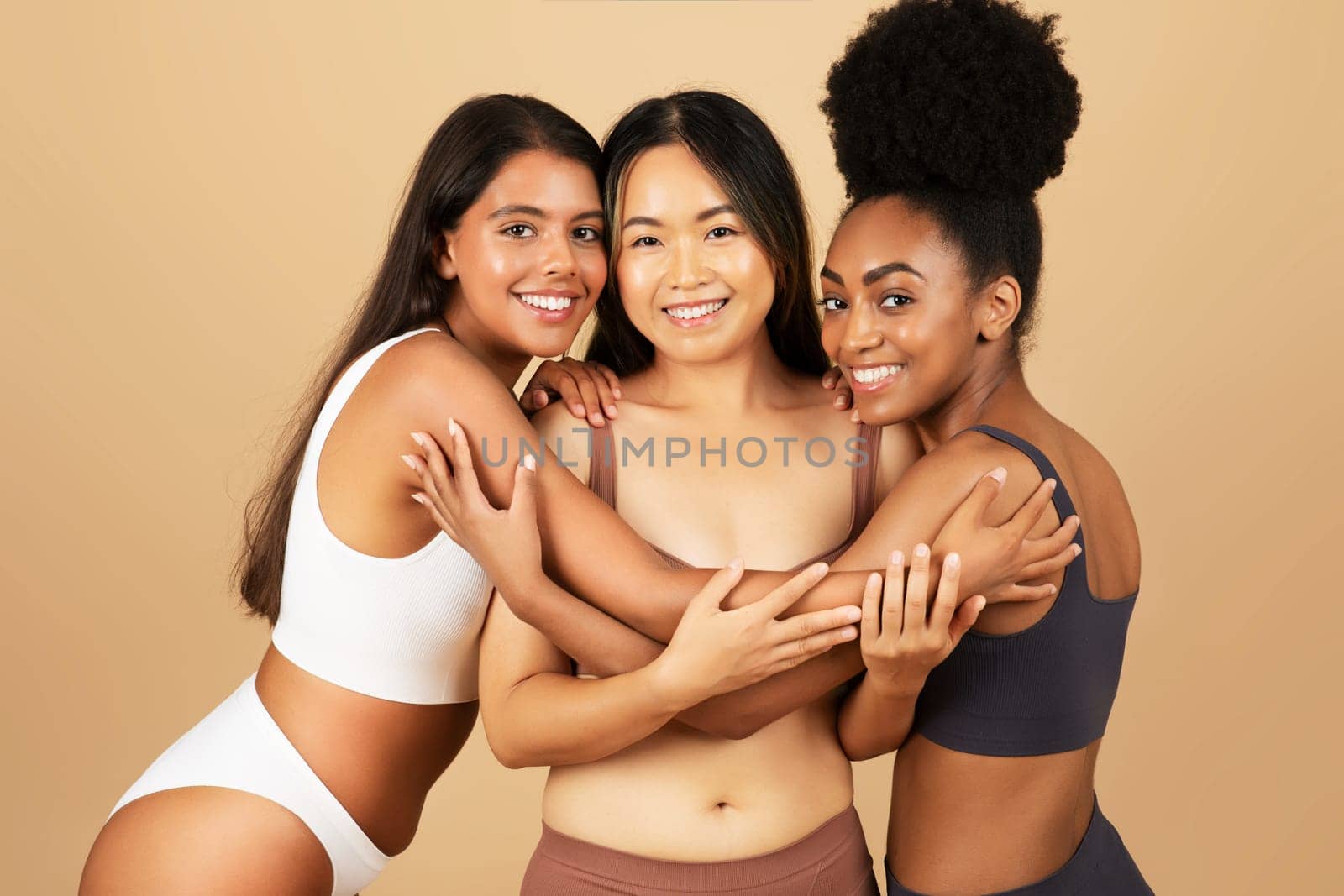 Three young women of different backgrounds smiling at camera, confidently pose and embrace in underwear, showcasing natural beauty on beige background.