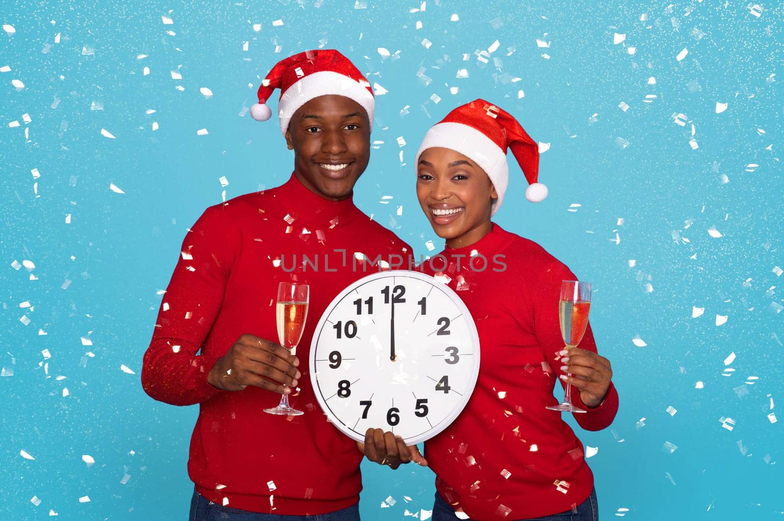 Happy African American Couple In Santa Hats Holding Champagne Glasses And Large Clock Amidst Falling Confetti On Blue Studio Background, Waiting Midnight Celebration And New Year's Eve Festivities