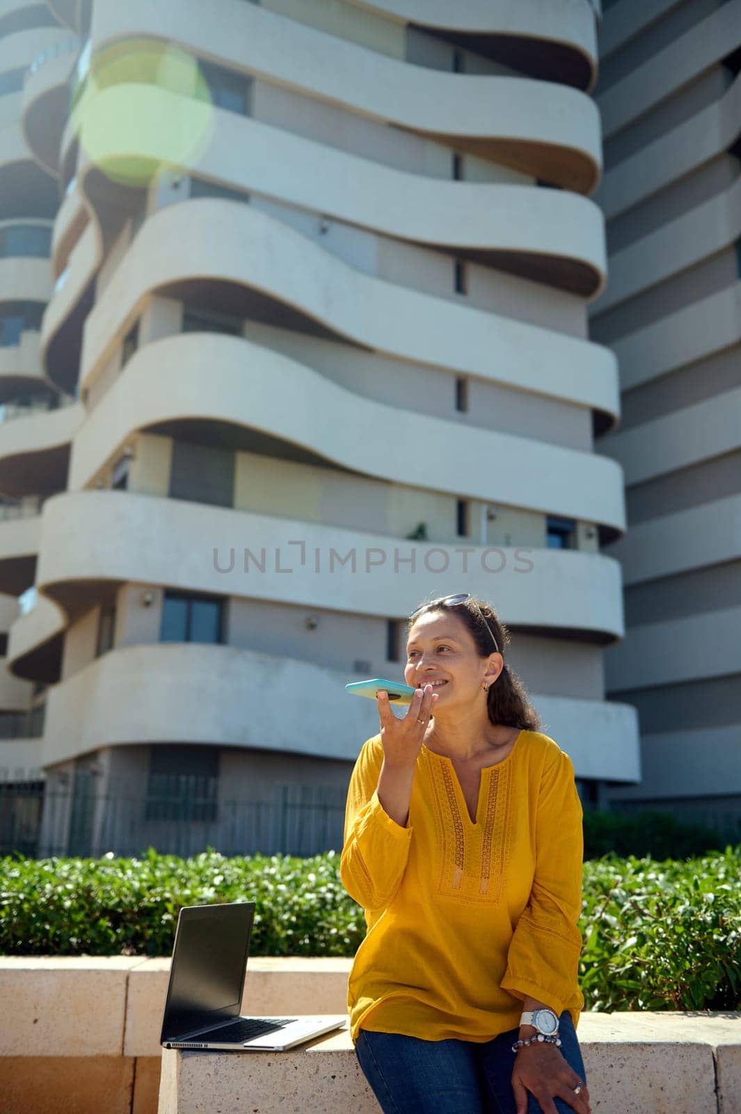 Vertical photography o a confident multi ethnic happy business woman using smart mobile phone, recording a voice message, remotely working against high rise buildings background. People. Business