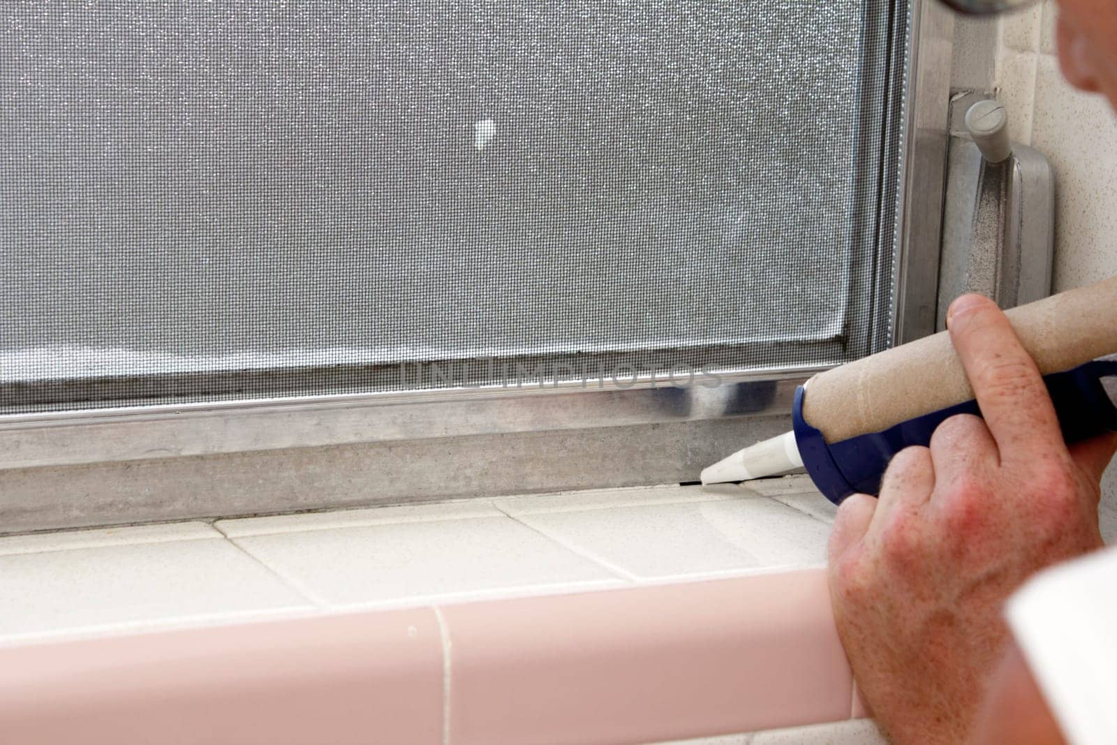 Caucasian male hand holding a blue caulking gun about to caulk a crack between an interior gray aluminum metal window frame and a speckled off white tile base of an old bathroom window sill in the day.