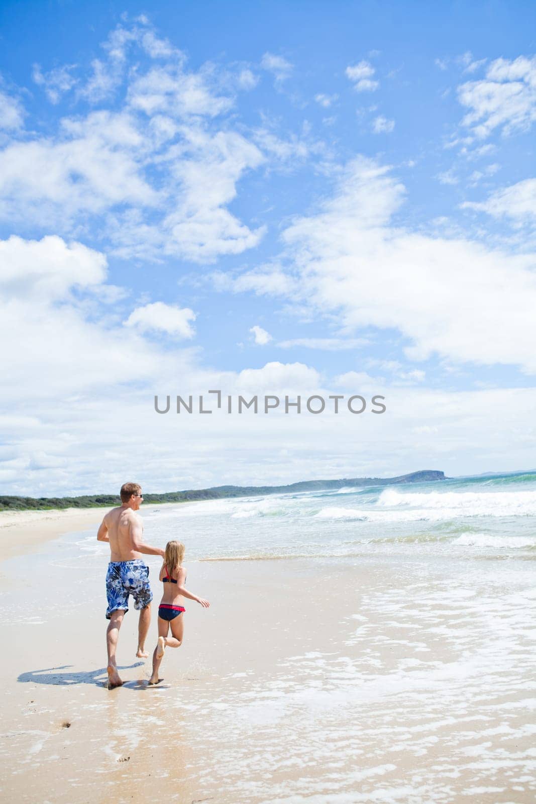 Father and young daughter holding hands and running along beach at edge of water