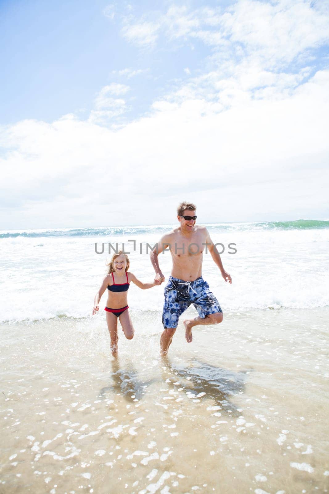 Happy smiling father and young daughter holding hands and running towards beach away from waves