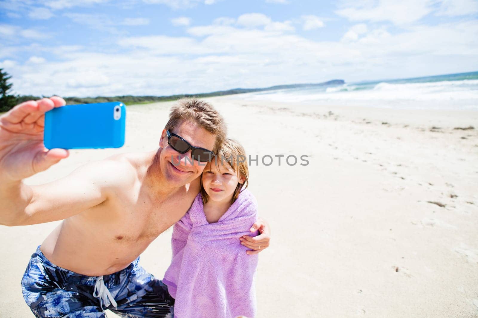 Smiling father taking selfie photograph with young daughter at beach using mobile phone