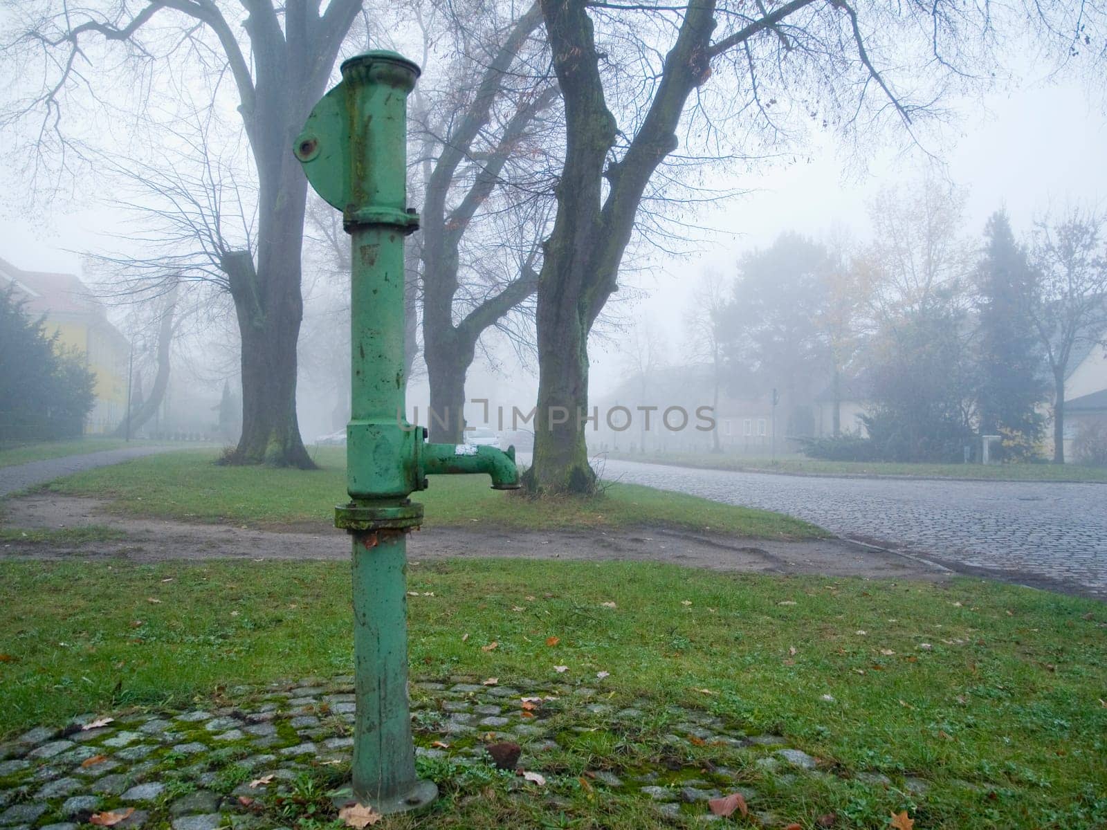 old green water pump on a village square in foggy November weather