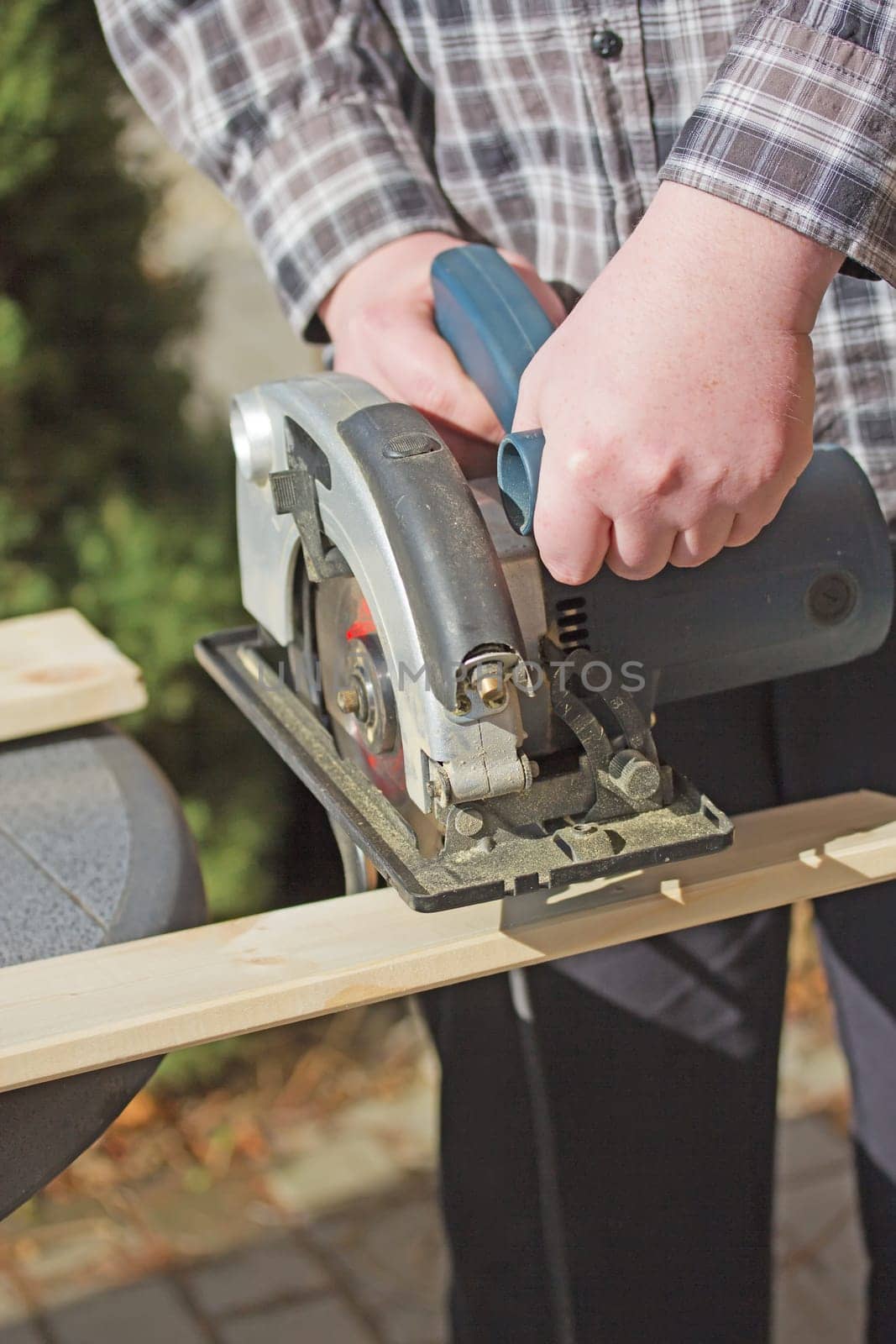 Closeup view of a man in a checkered shirt that is cutting wooden board by electric jigsaw outdoors.