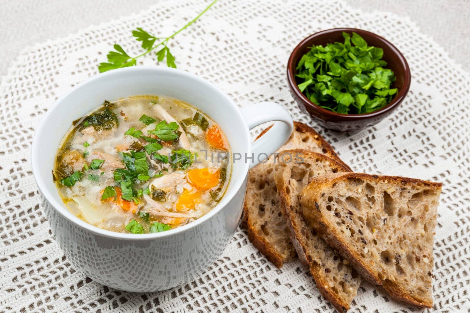 Cup of hot chicken rice soup served with bread and parsley on crochet tablecloth