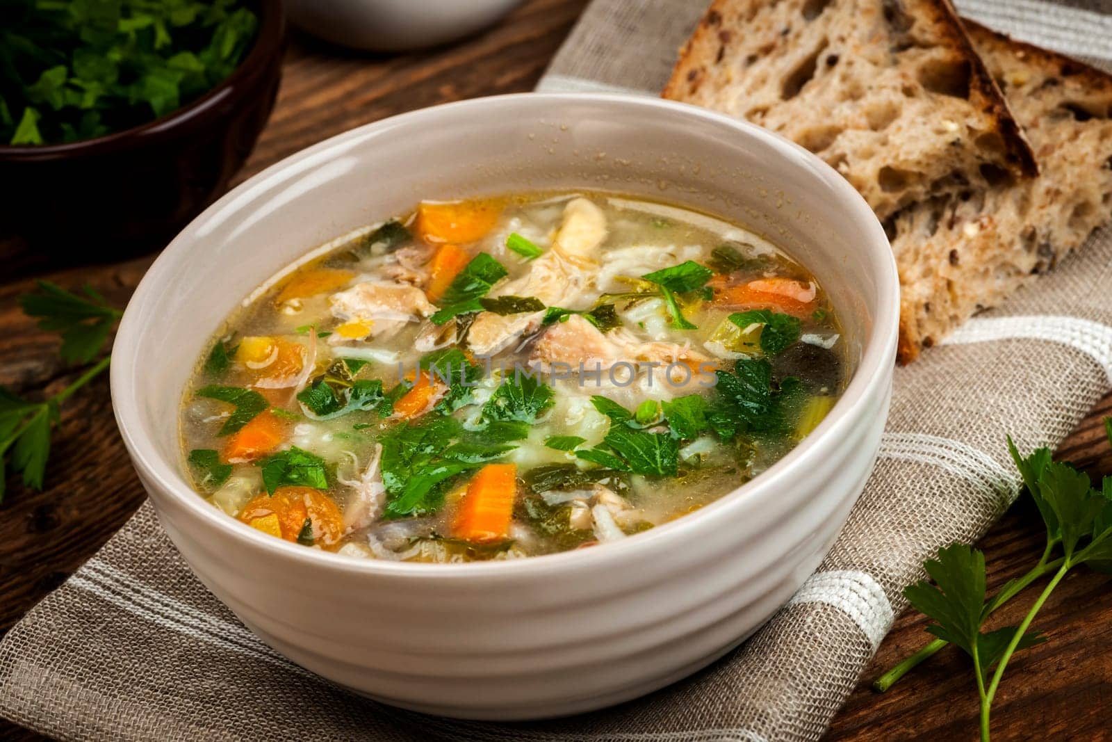 Chicken rice soup with vegetables in bowl and bread on rustic table