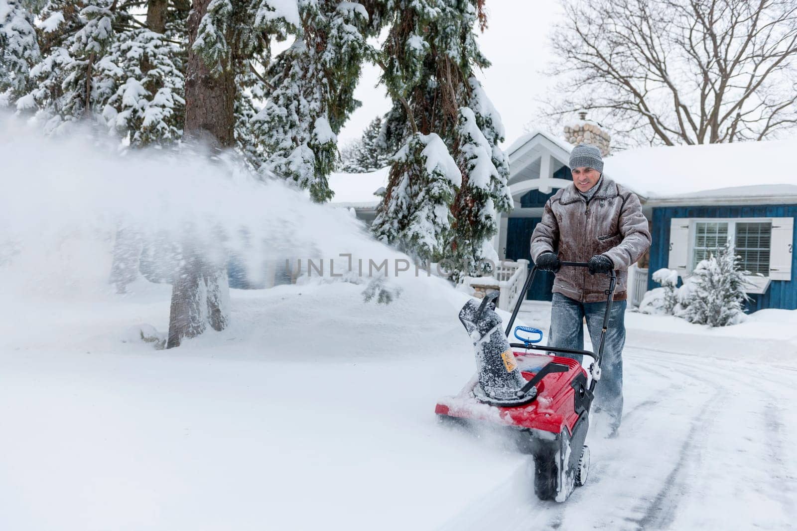 Man using snowblower to clear deep snow on driveway near residential house after heavy snowfall.
