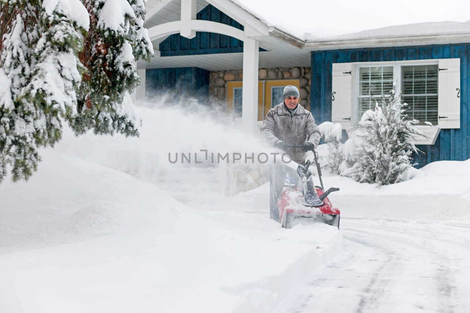 Man using snowblower to clear deep snow on driveway near residential house after heavy snowfall.