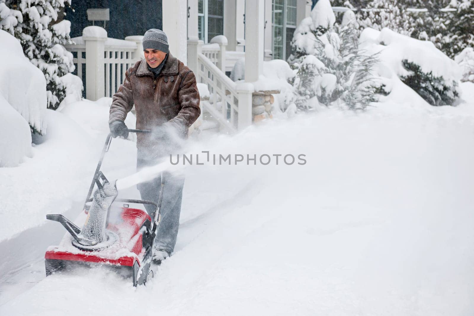Man using snowblower to clear deep snow on driveway near residential house after heavy snowfall.
