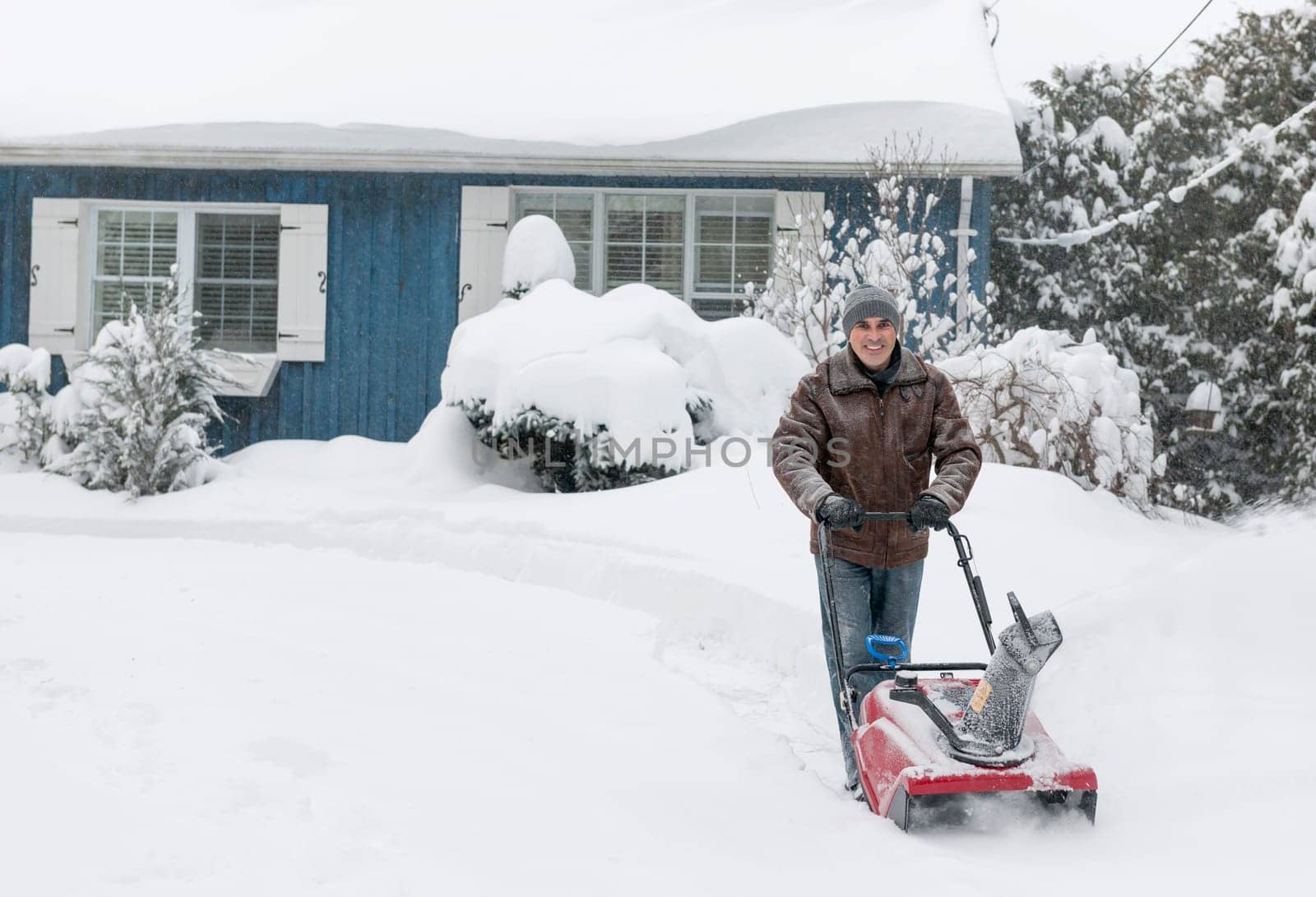 Man using snowblower to clear deep snow on driveway near residential house after heavy snowfall.