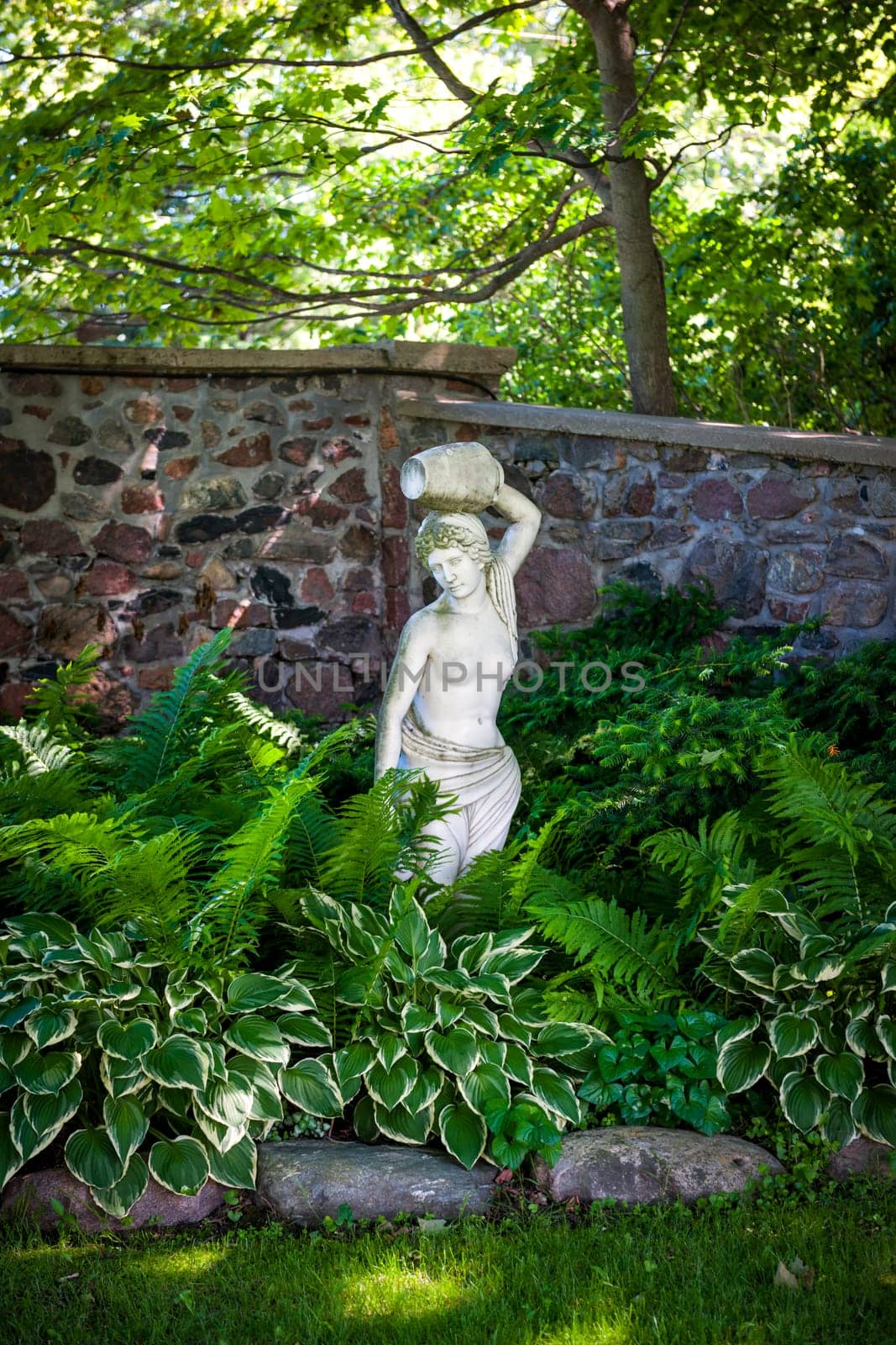 Lush green summer garden with perennial plants and statue near stone wall