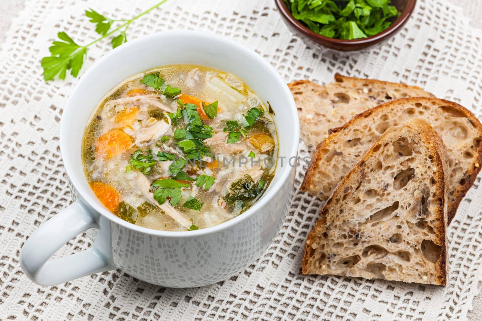 Cup of hot chicken rice soup served with bread and parsley on crochet tablecloth