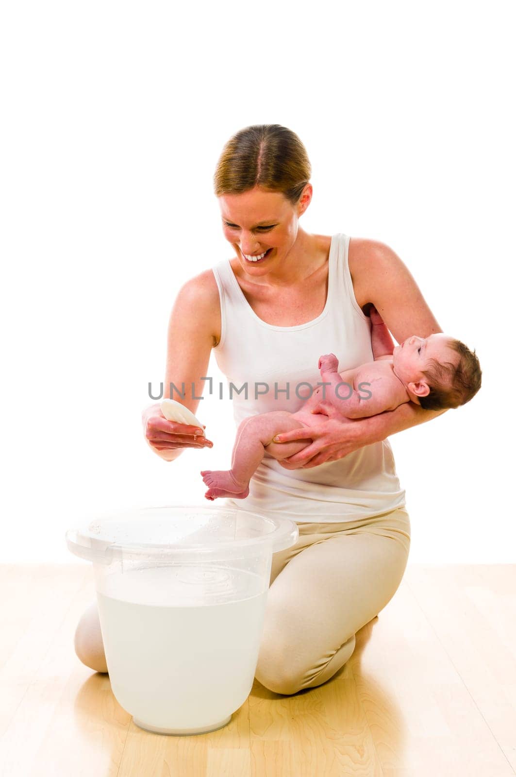 Adult mother wearing bright casual clothes, she kneels with her newborn baby on the floor, in front of a bathing bucket. In her hand she holds a thermometer and checked the termperature of the bath water. Isolated against white background.