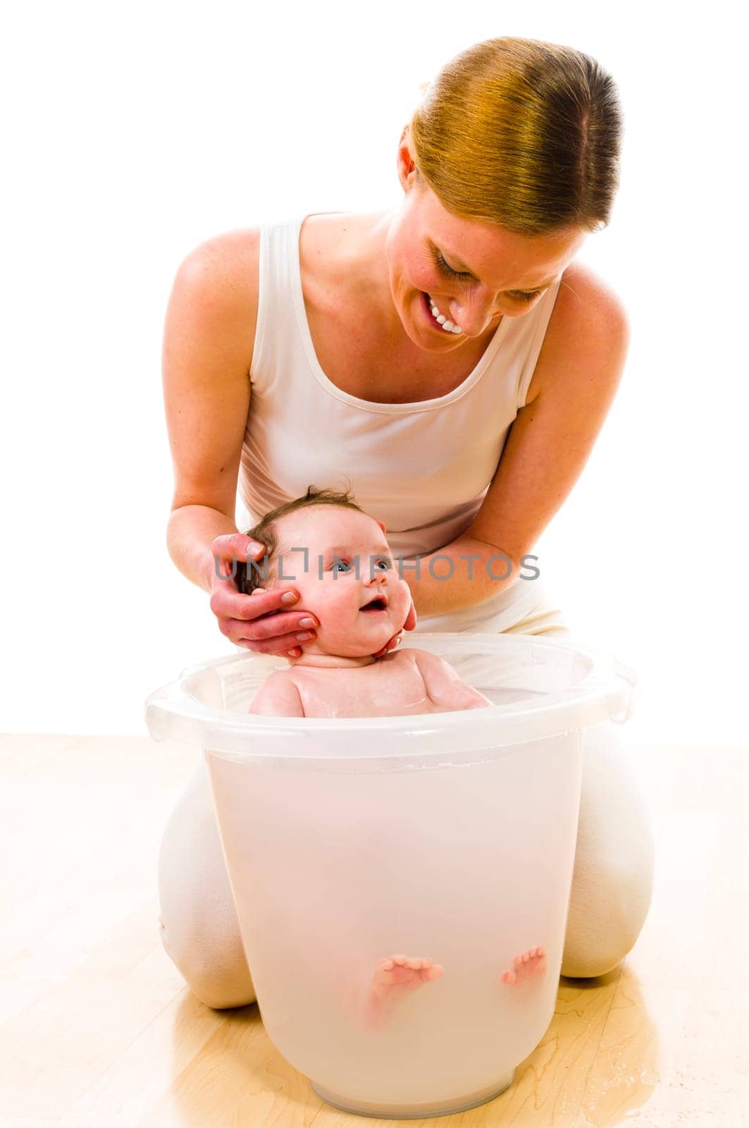 Adult mother wearing bright casual clothes, she kneels on the floor and bathes her newborn baby in the bath bucket. Carefully, she keeps the baby's head. Isolated against white background.