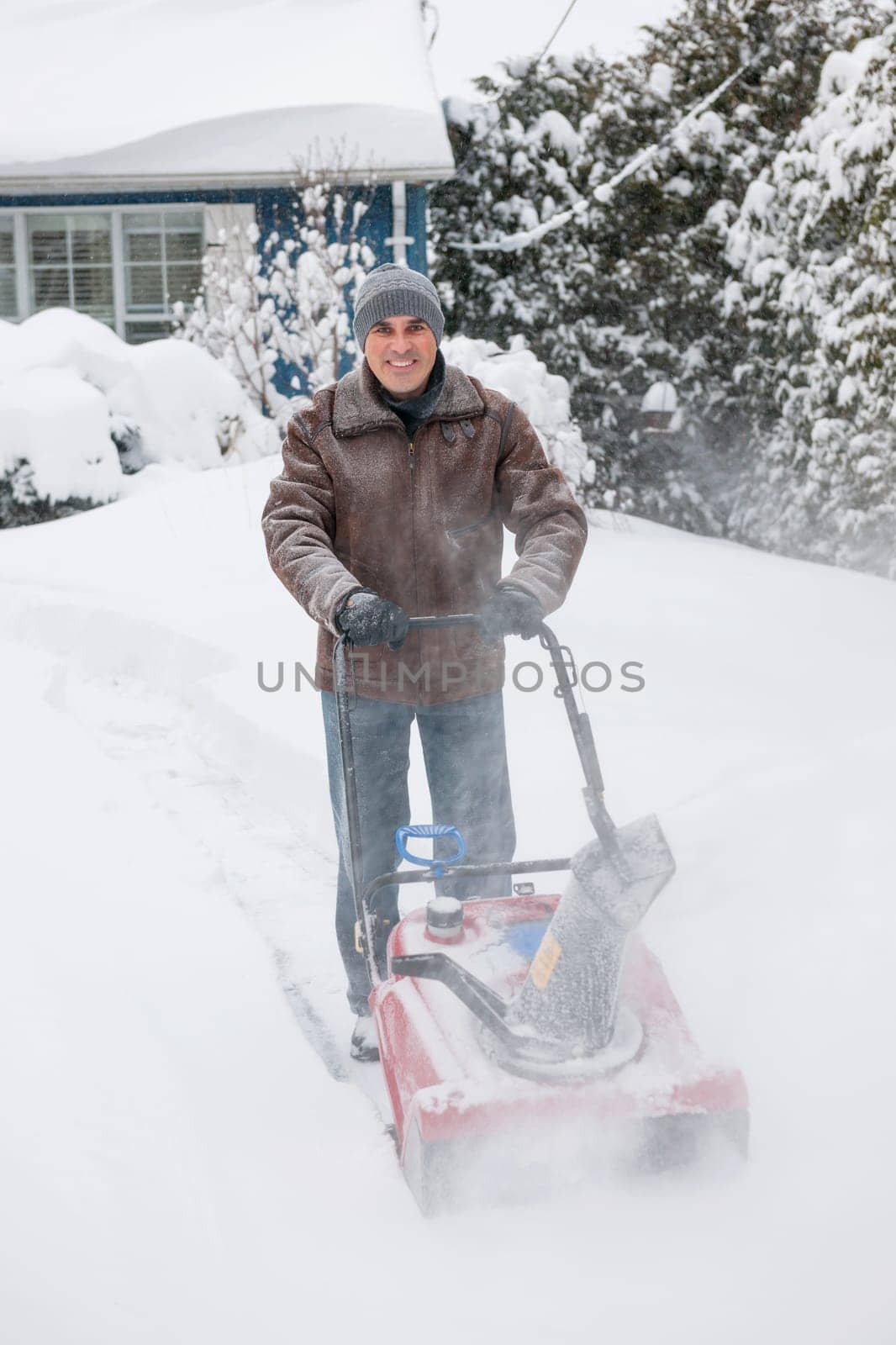 Man using snowblower to clear deep snow on driveway near residential house after heavy snowfall.