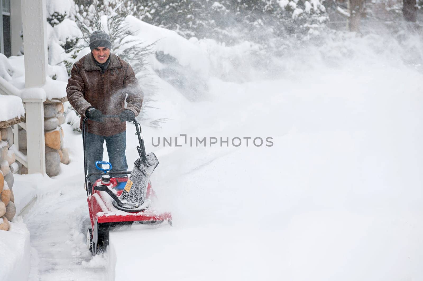 Man using snowblower to clear deep snow on driveway near residential house after heavy snowfall.