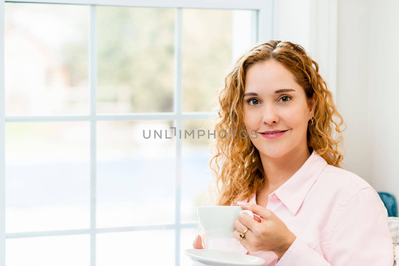 Smiling caucasian woman relaxing by window holding cup of coffee