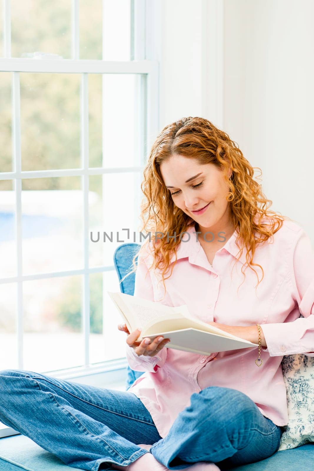 Smiling caucasian woman reading book by window at home