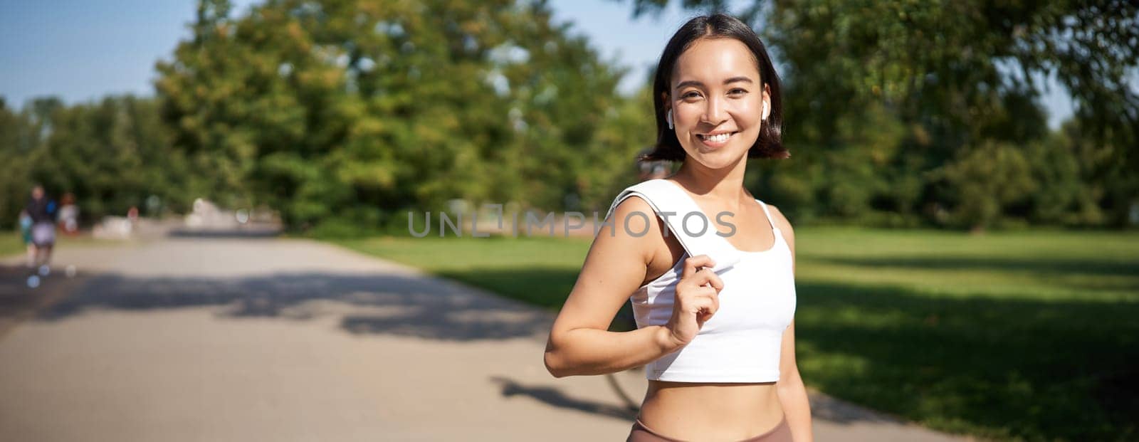 Smiling asian fitness girl holding towel on shoulder, workout in park, sweating after training exercises outdoors by Benzoix
