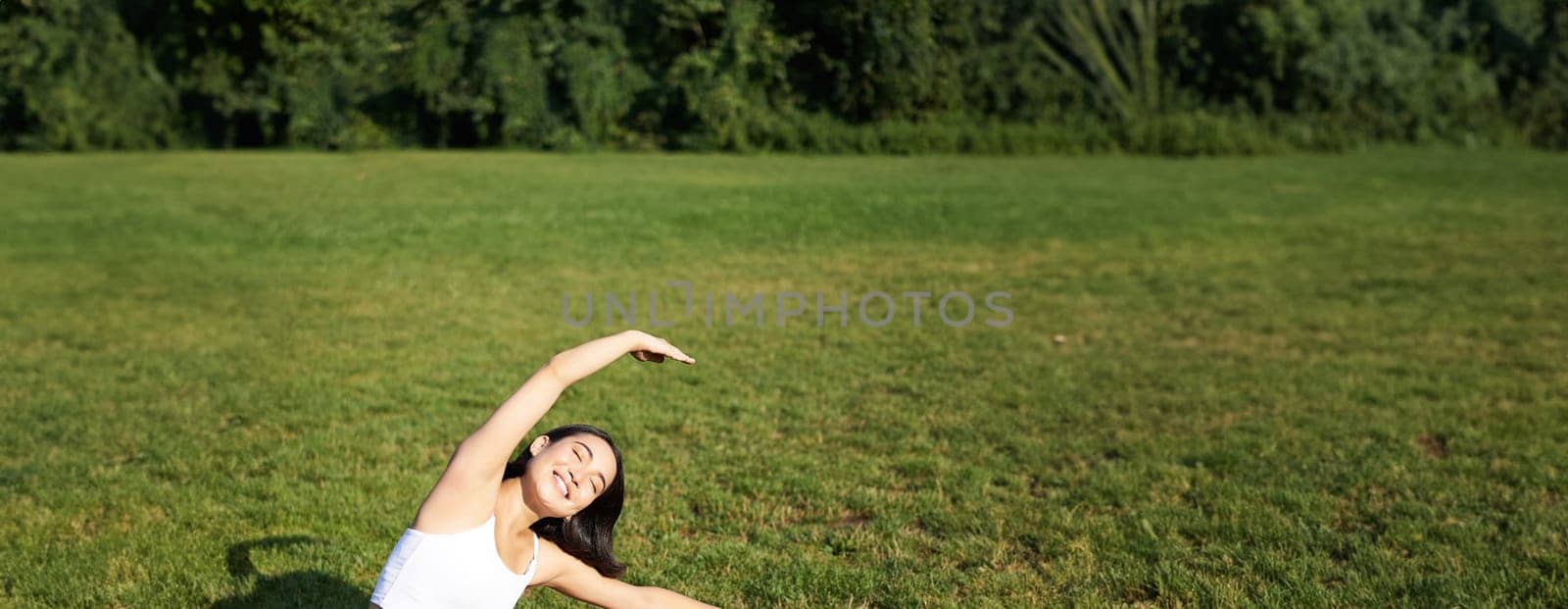 Young woman does yoga on lawn in park, stretching on fitness mat, wellbeing concept.