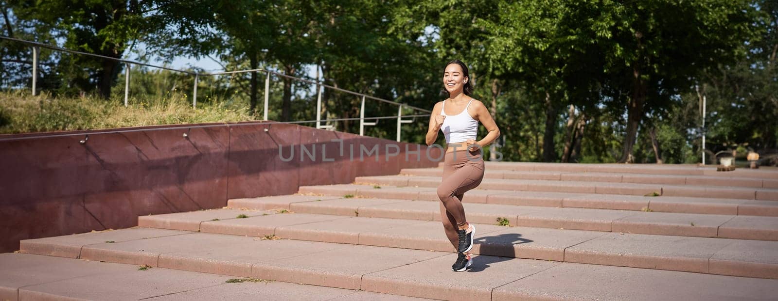 Healthy fitness girl running outdoors on street, wearing uniform, jogging on fresh air and listening music in wireless headphones.