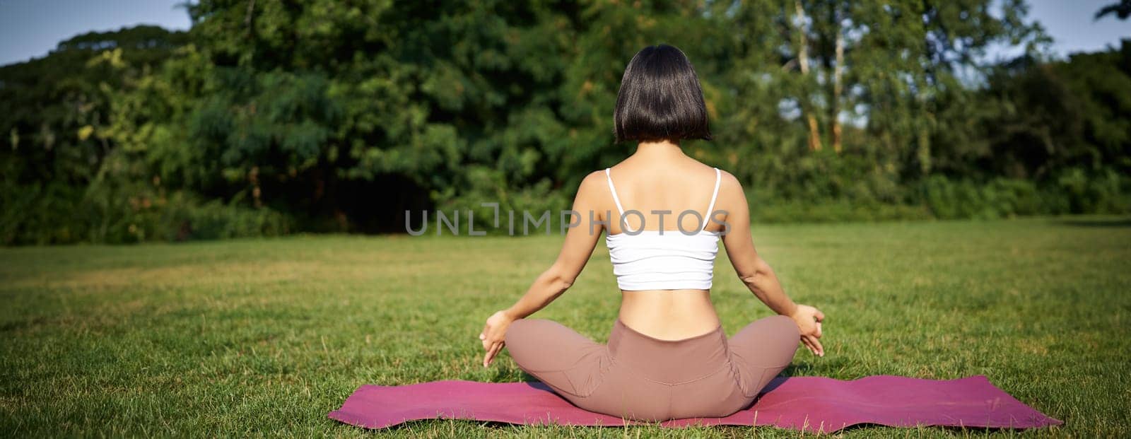 Rear view of woman silhouette doing yoga, sitting on fitness mat and meditating on green lawn by Benzoix