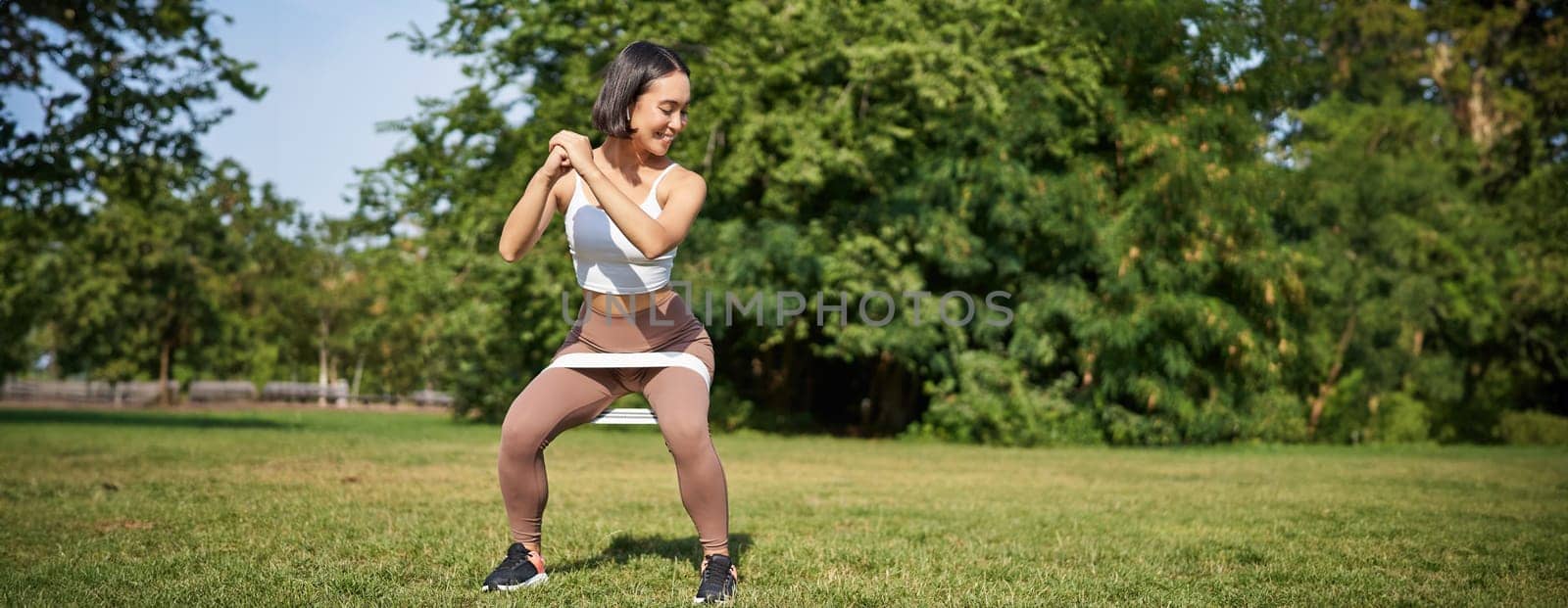 Woman using stretching band, resistance fitness rope for workout in park, doing squats, training on fresh air by Benzoix