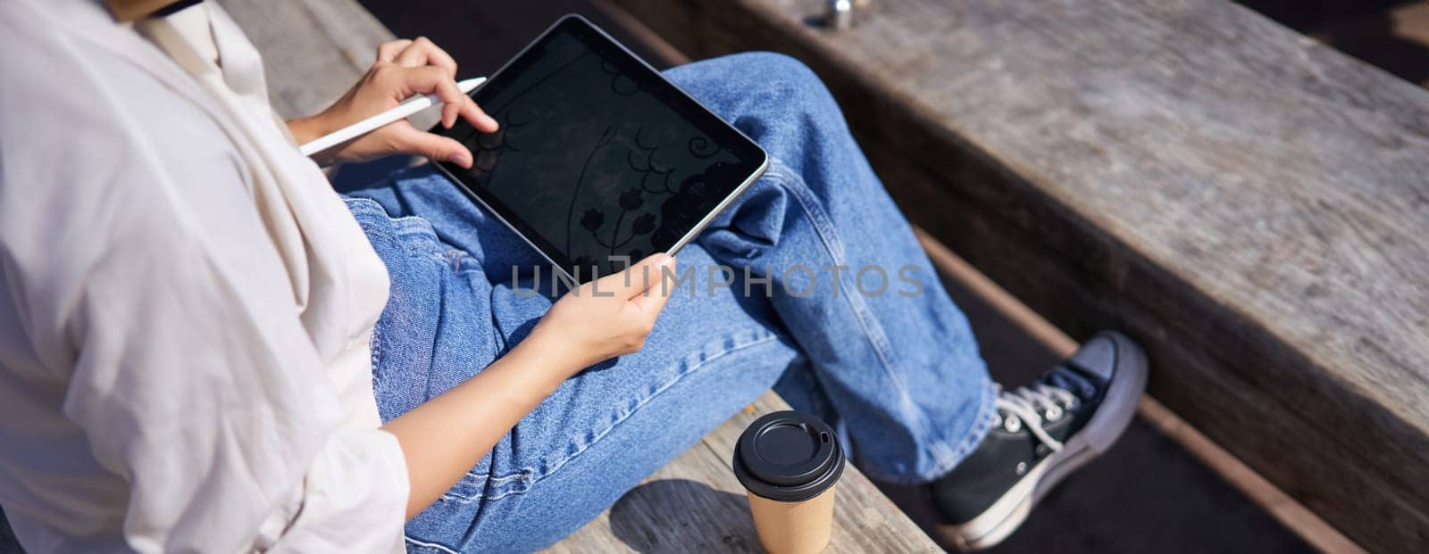 Cropped shot of female hands drawing on digital graphic tablet with pen, sitting with cup of coffee outdoors on bench.
