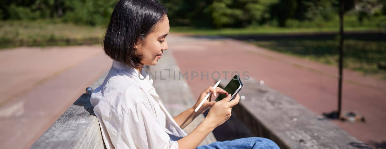 Asian girl, drawing in park, sitting on street on sunny day with digital graphic tablet and pen, smiling happily.