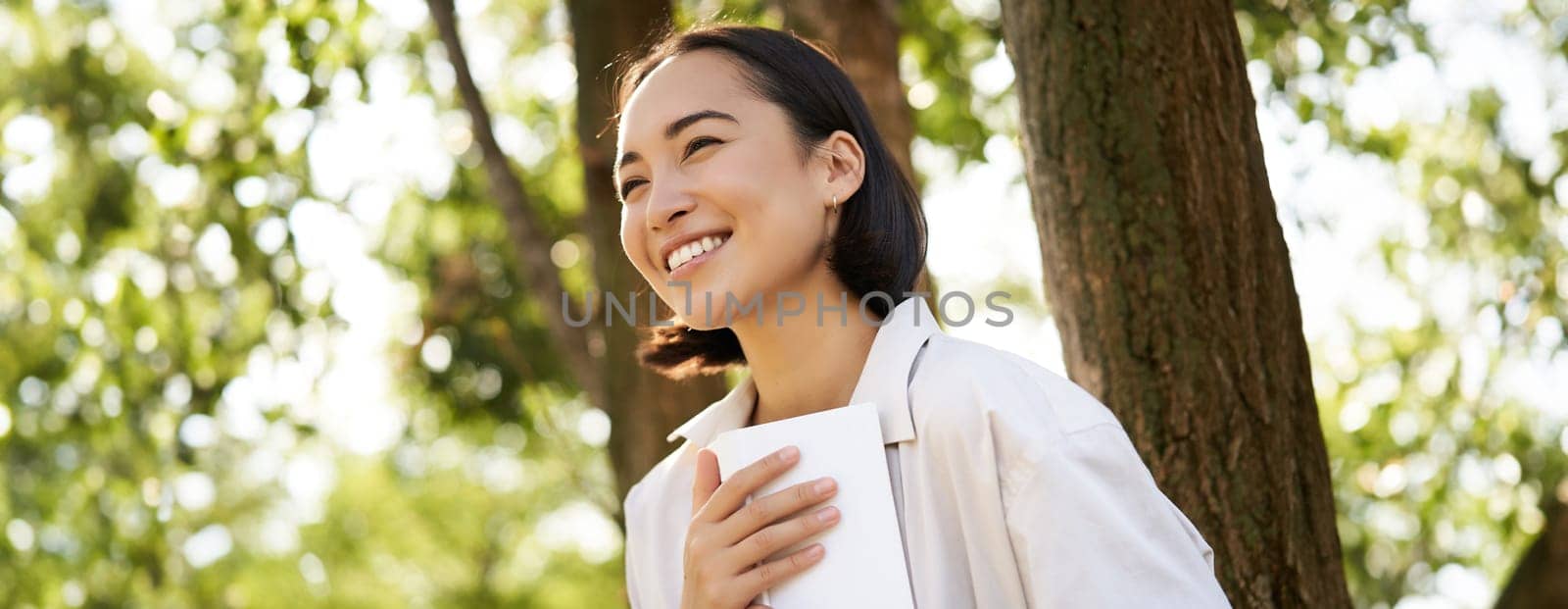 Romantic smiling girl reading book in park or foret, sitting under tree shade on sunny day, relaxing on fresh air surrounded by nature.
