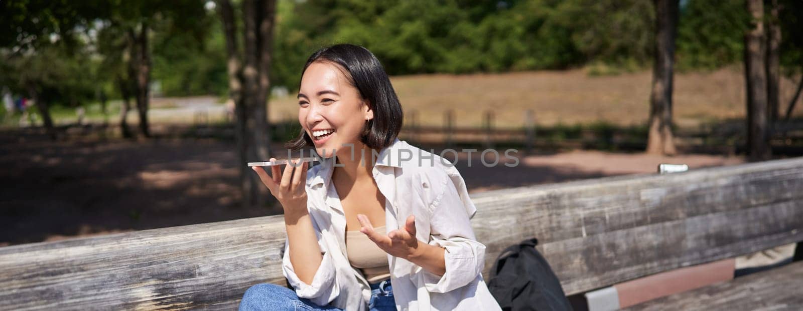 Portrait of happy asian girl records voice message, talking via speakerphone on smartphone, sitting relaxed on bench in park on sunny day by Benzoix
