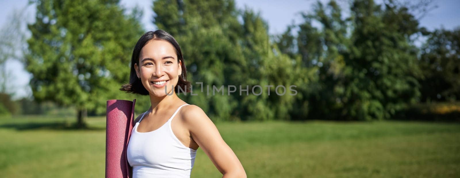 Smiling fitness girl with rubber mat, stands in park wearing uniform for workout and sport activities, does yoga outdoors on lawn by Benzoix