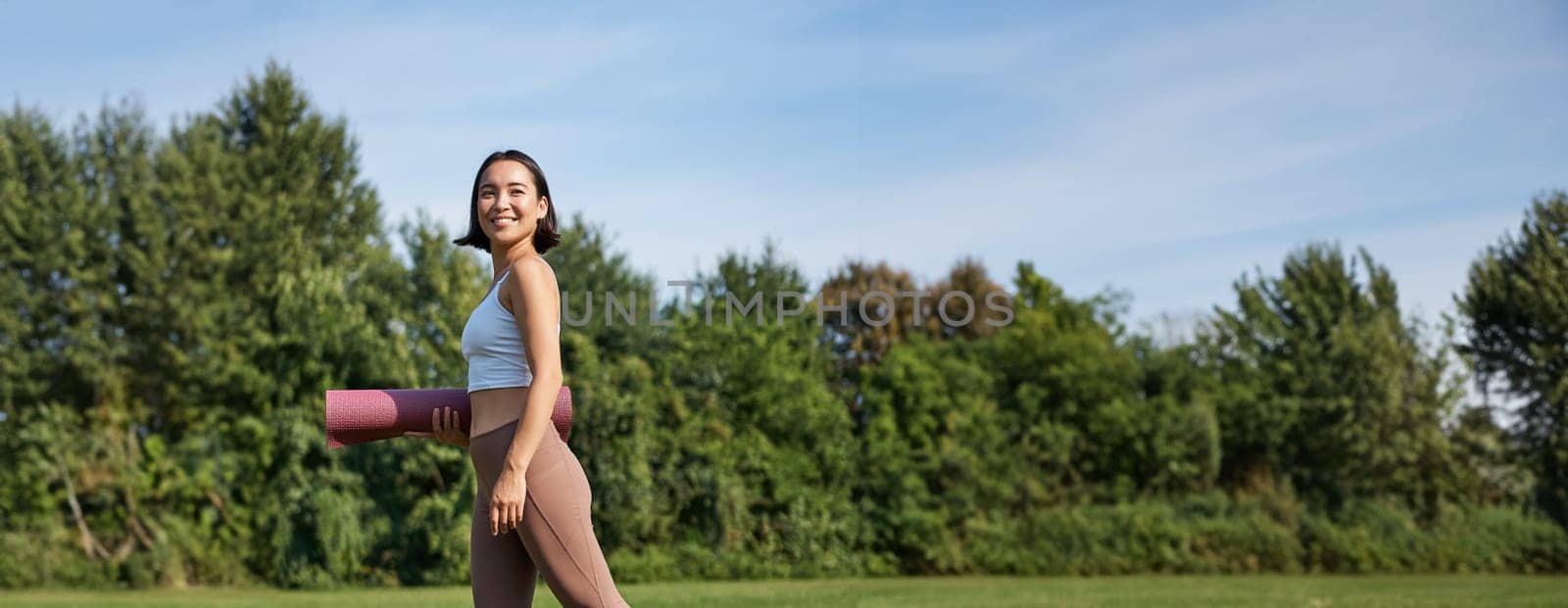 Vertical shot of stylish fitness woman with rubber mat, posing on lawn in park, doing workout on fresh air.