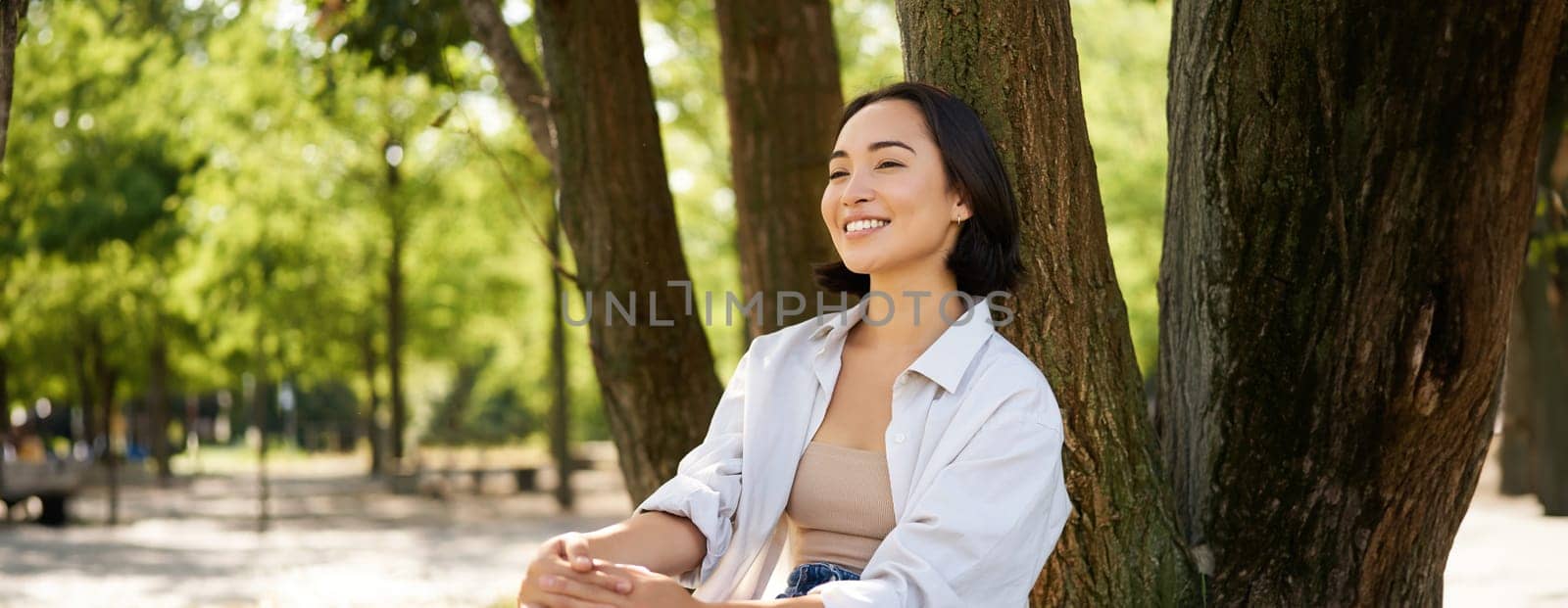 Portrait of asian girl relaxing, leaning on tree and resting in park under shade, smiling and enjoying the walk outdoors by Benzoix