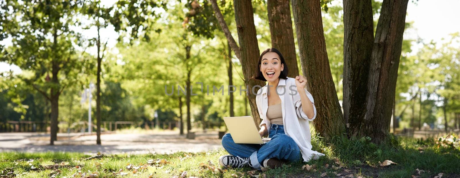 Portrait of asian young woman, student doing homework, working in park, sitting beside tree with laptop and showing okay sign, approve smth by Benzoix
