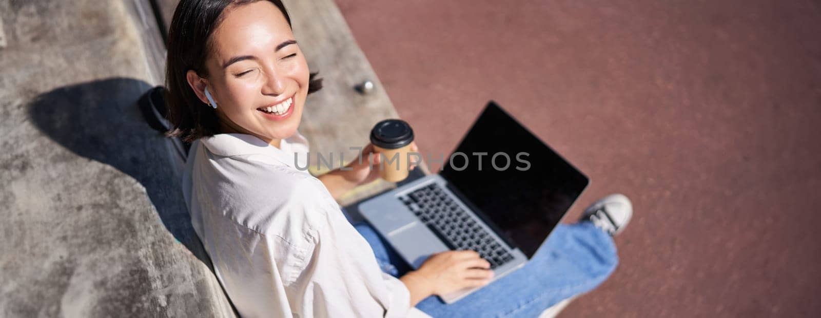 Beautiful young asian female student, sitting with laptop on bench in park, drinking takeaway coffee and listening music, working remotely, studying outdoors by Benzoix