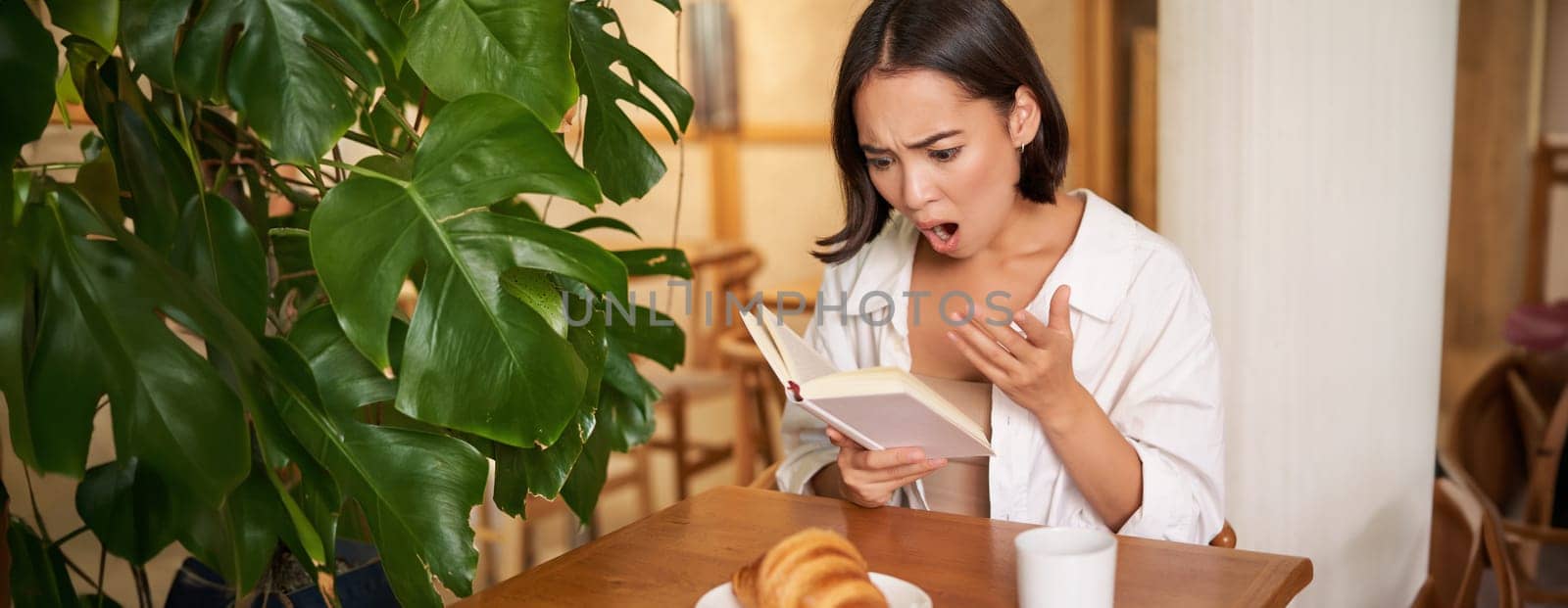 Portrait of asian woman looking shocked at book pages, reading something interesting, concentrating, sitting in cafe with coffee and croissant by Benzoix