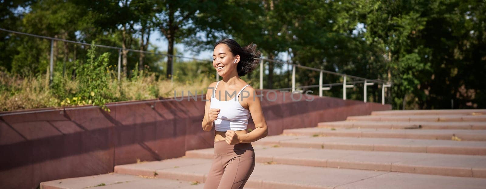 Healthy fitness girl running outdoors on street, wearing uniform, jogging on fresh air and listening music in wireless headphones by Benzoix
