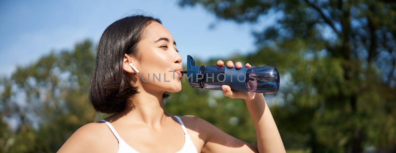 Happy asian sportswoman, runner drinks water from bottle while running, workout on fresh air in park by Benzoix