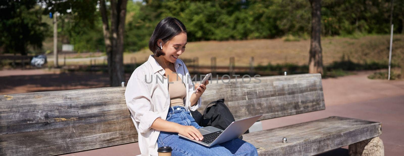 Young asian girl talking on mobile phone, using laptop, working online from park, sitting on bench on sunny day with computer by Benzoix