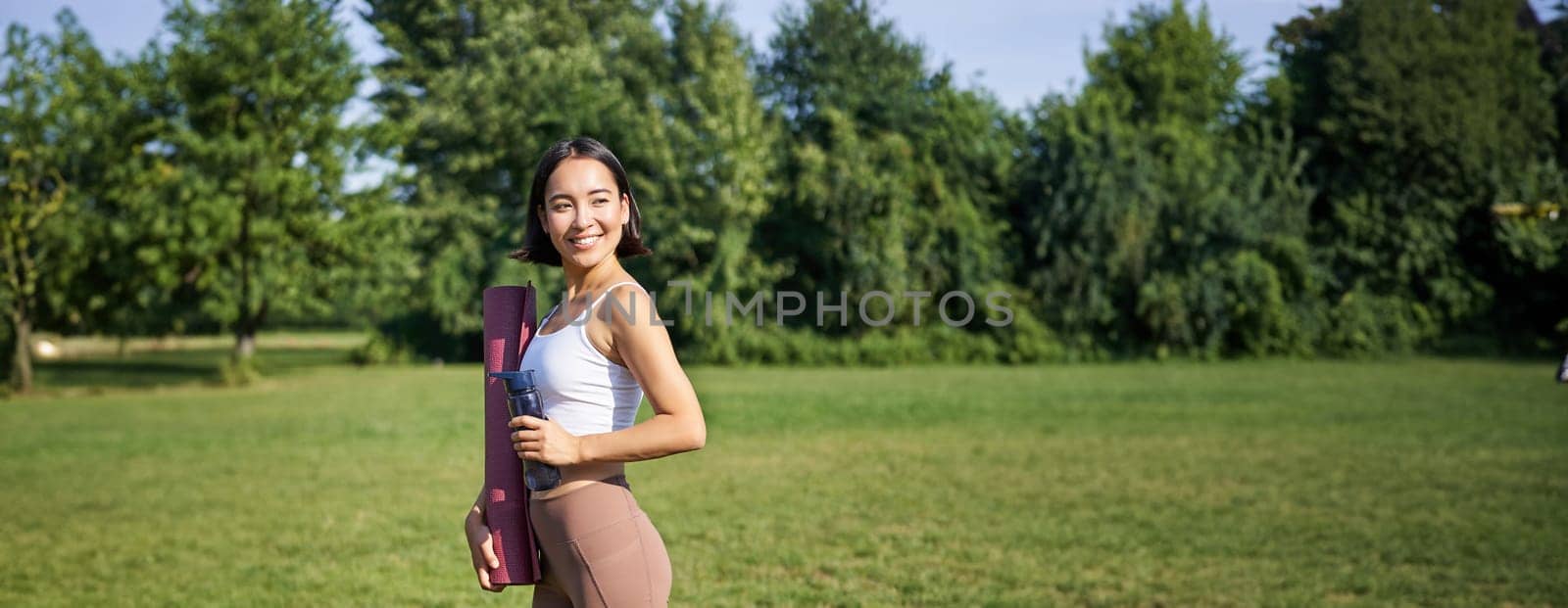 Vertical shot of fit and healthy asian woman posing in park, holding water bottle and yoga rubber mat for workout outdoors by Benzoix